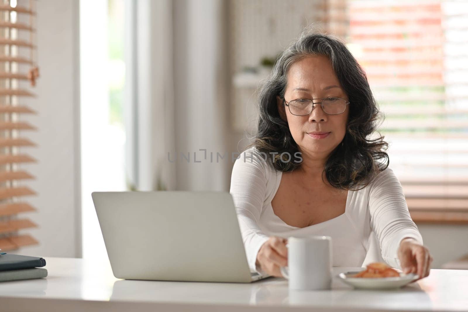 Happy asian senior woman having breakfast and using laptop on desk at home. Elderly lifestyle and technology concept.