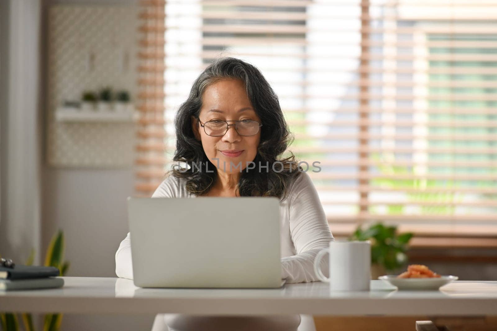 Contemporary senior female in glasses watching webinar or checking email on laptop computer at home.