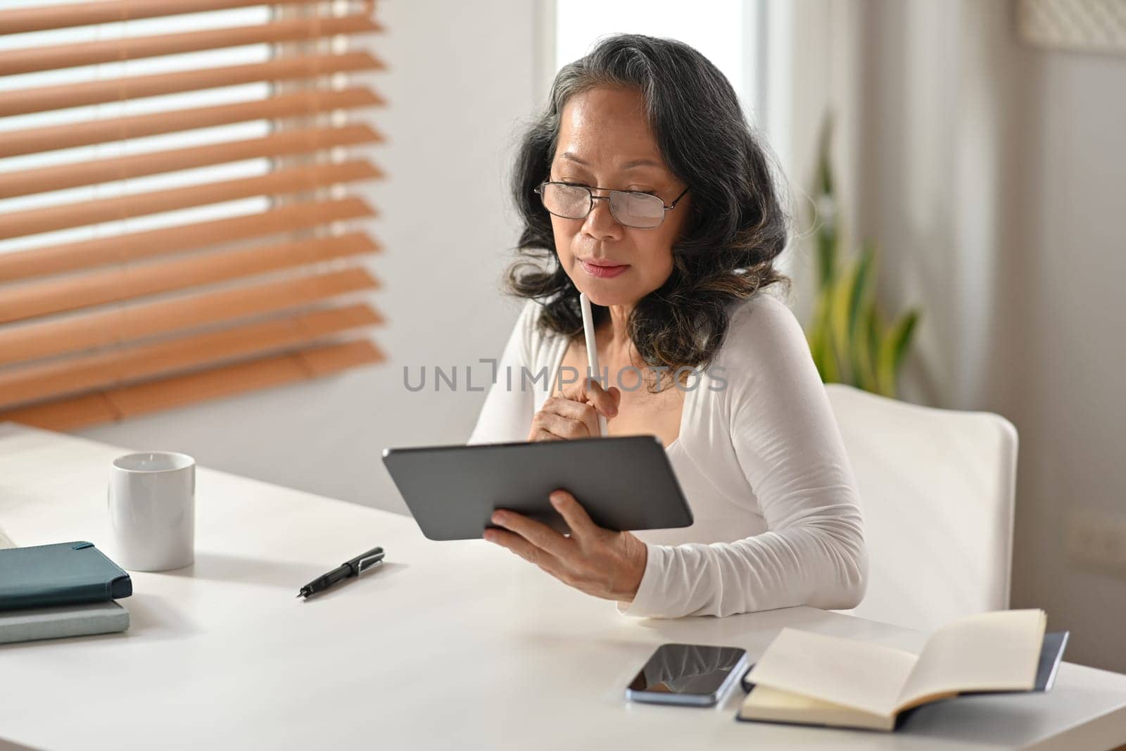 Smiling middle aged Asian woman sitting in cozy home browsing wireless Internet on digital tablet. Elderly technology concept.
