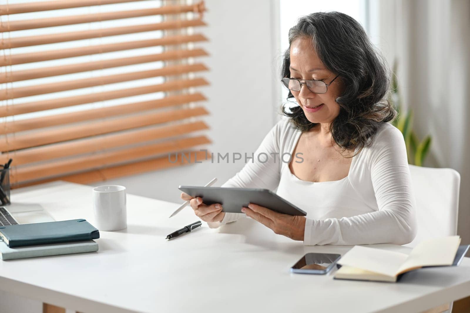 Image of happy middle age woman sitting in home office communicating in social media, resting at workplace afterwork by prathanchorruangsak