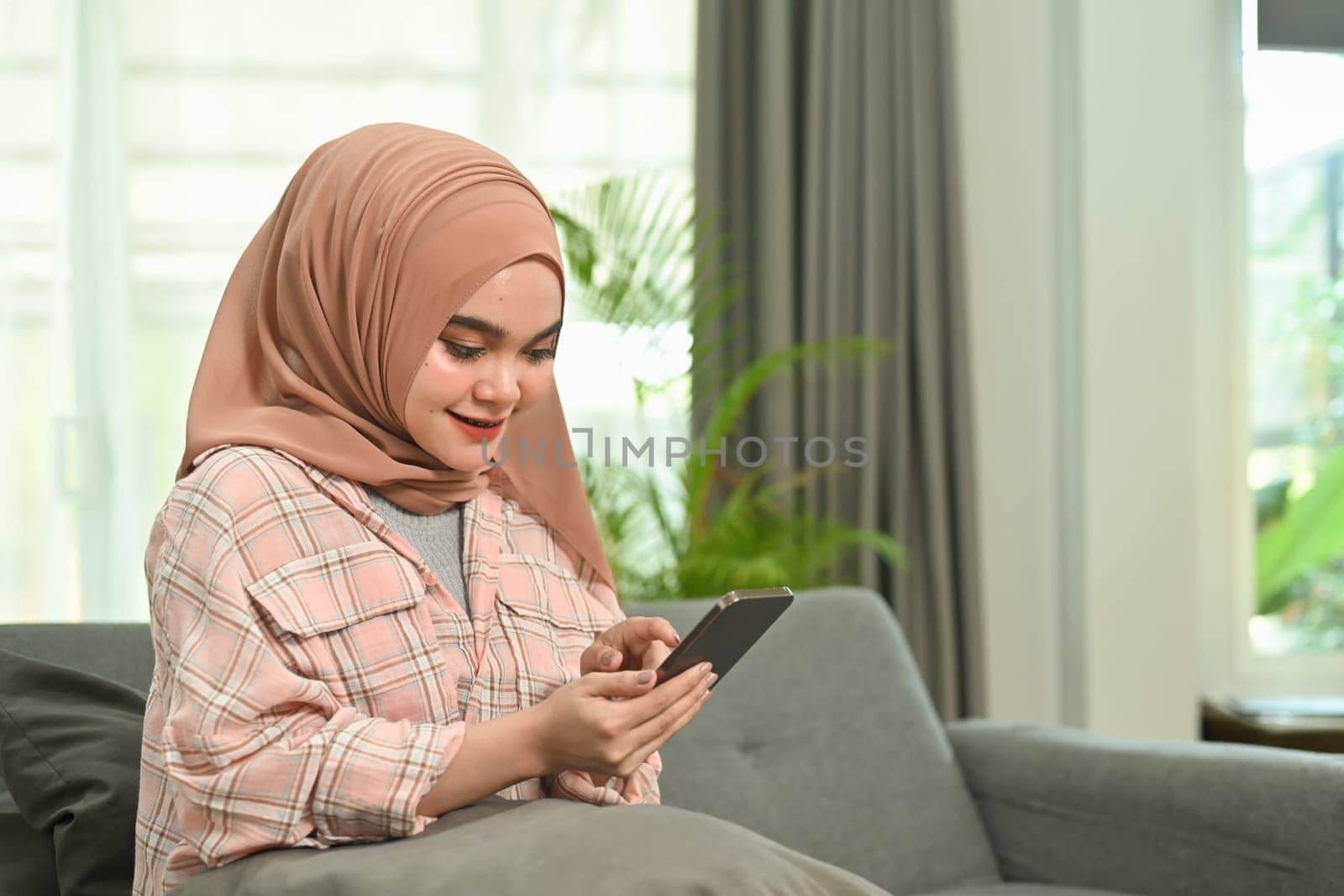Beautiful Muslim woman sitting on couch texting messaging, browsing wireless internet on smartphone.