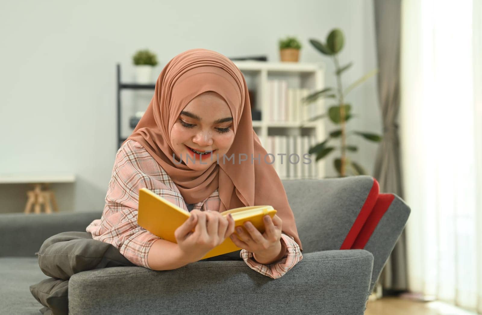 Portrait of joyful muslim woman in hijab lying on comfortable couch at home and reading book.
