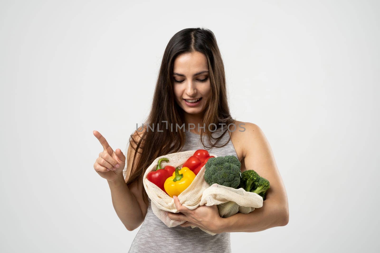 Smiling brunette girl is holding mesh shopping bag with vegetables, greens without plastic bags. Zero waste, plastic free Eco friendly concept. Sustainable lifestyle