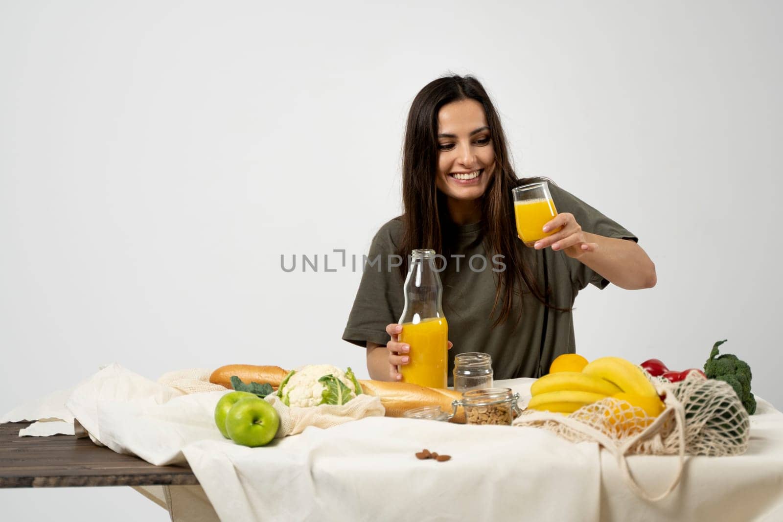 Woman in green t-shirt pouring a juice from a glass bottle in a glass over a table with mesh eco bag, healthy vegan vegetables, fruits, bread, snacks. Zero waste concept. by vovsht