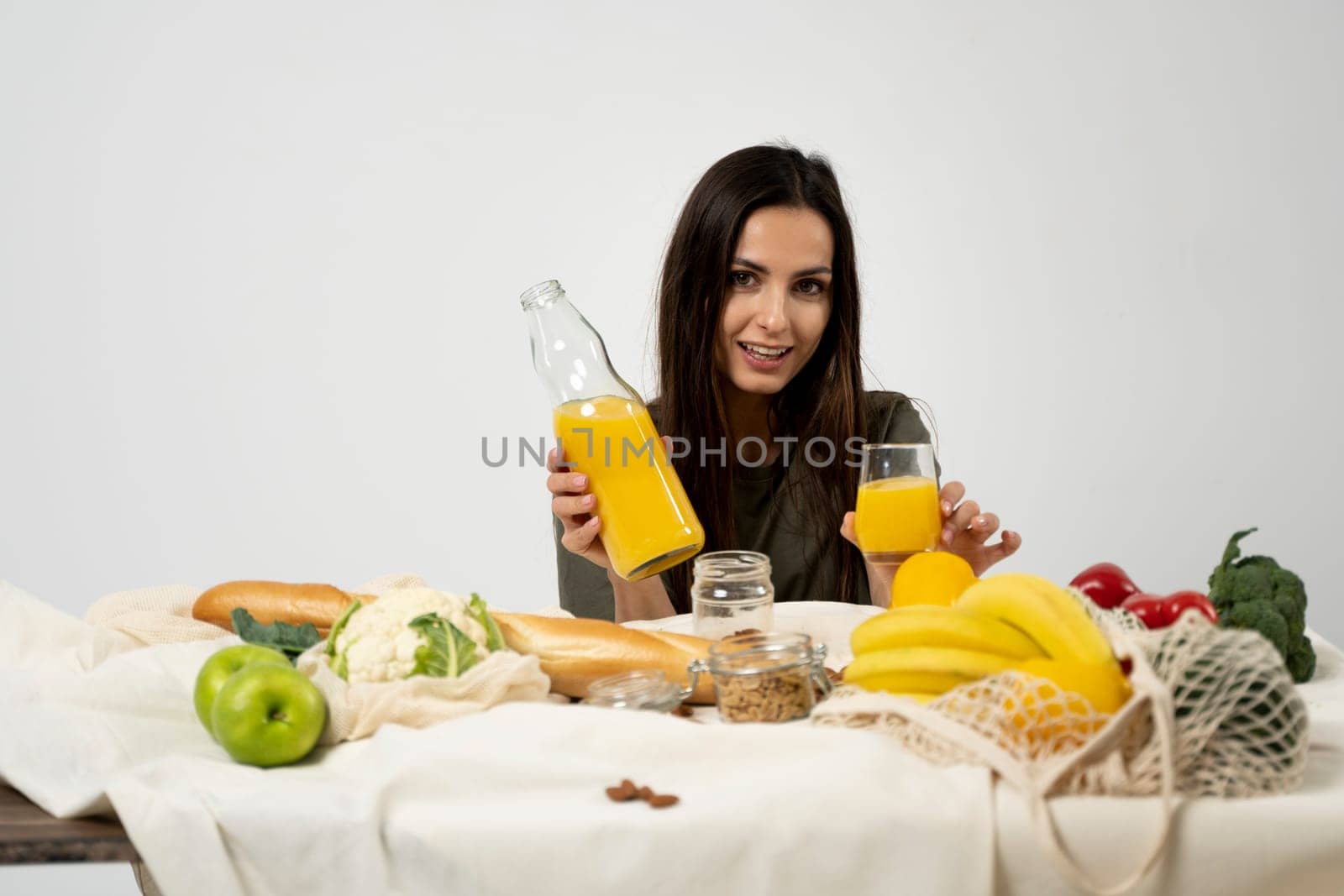 Woman in green t-shirt drink a juice over a table with mesh eco bag, healthy vegan vegetables, fruits, bread, snacks. Zero waste concept. by vovsht