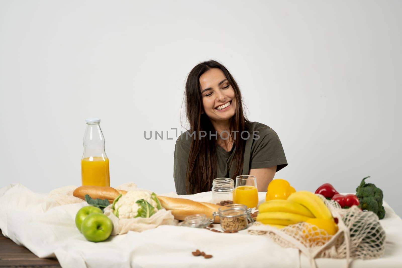 Happy woman in green t-shirt unpacking shopping mesh eco bag with healthy vegetables, fruits, bread, snacks on the kitchen at home. Healthy eating vegetarian concept. by vovsht