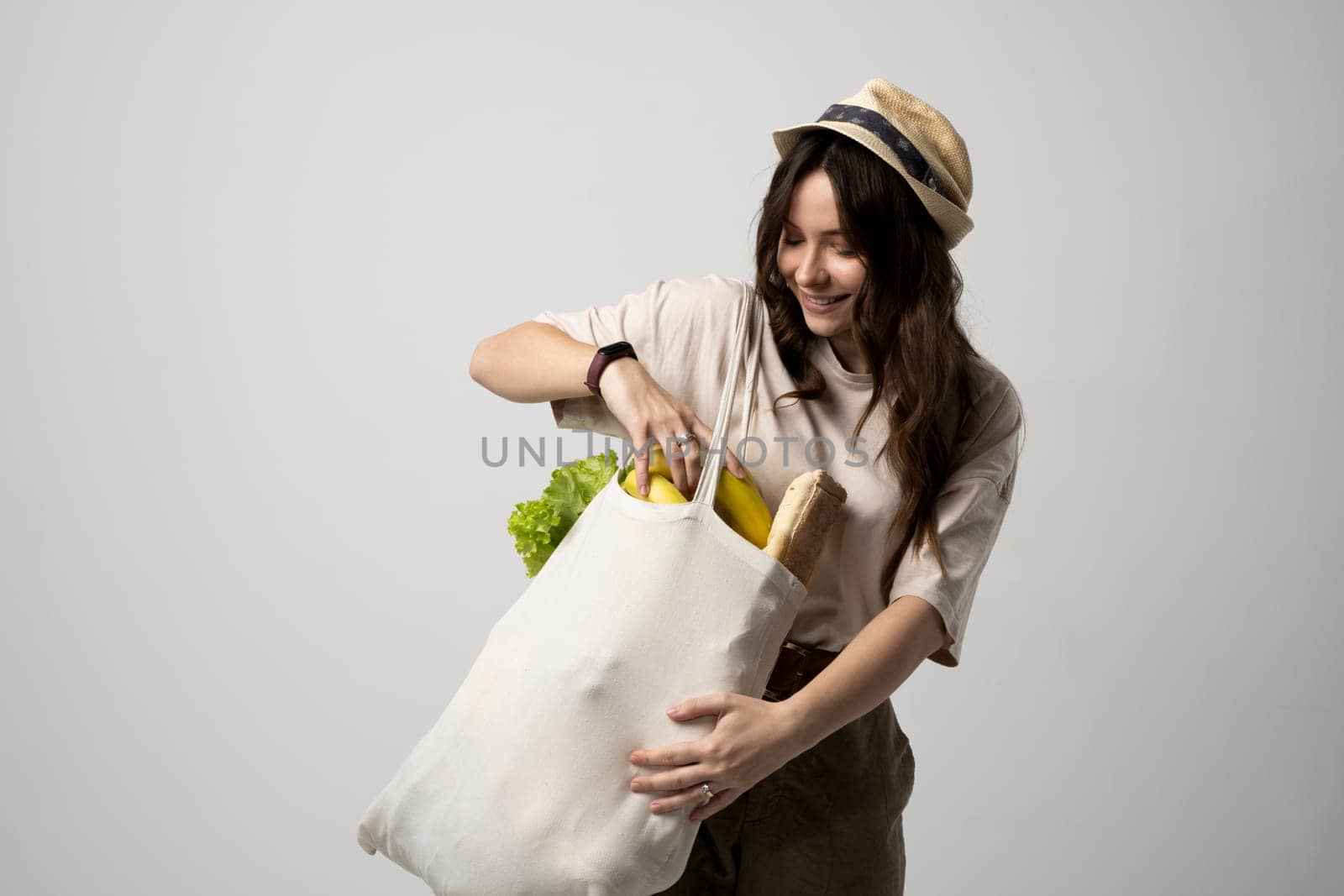 Smiling woman in a t-shirt and a hat holding reusable cotton shopping bag with vegetables, bread and greens. Concept of no plastic, zero waste, plastic free, sustainable lifestyle. by vovsht