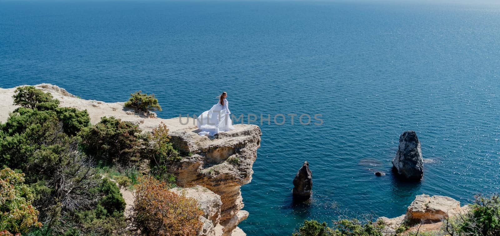 Woman in a white dress on the sea. Side view Young beautiful sensual woman in white long dress posing on a rock high above the sea at sunset. Girl in nature against the blue sky by Matiunina