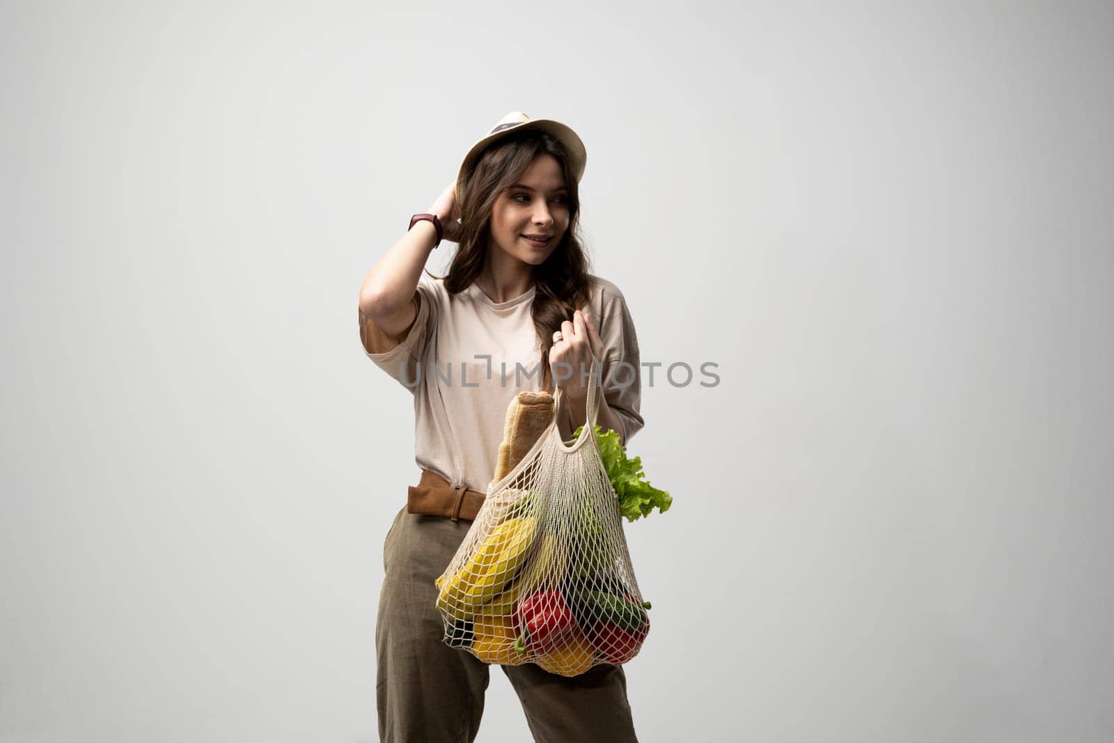 Eco friendly positive young woman in beige oversize t-shirt holding reusable mesh cotton eco bags for shopping with groceries on white background
