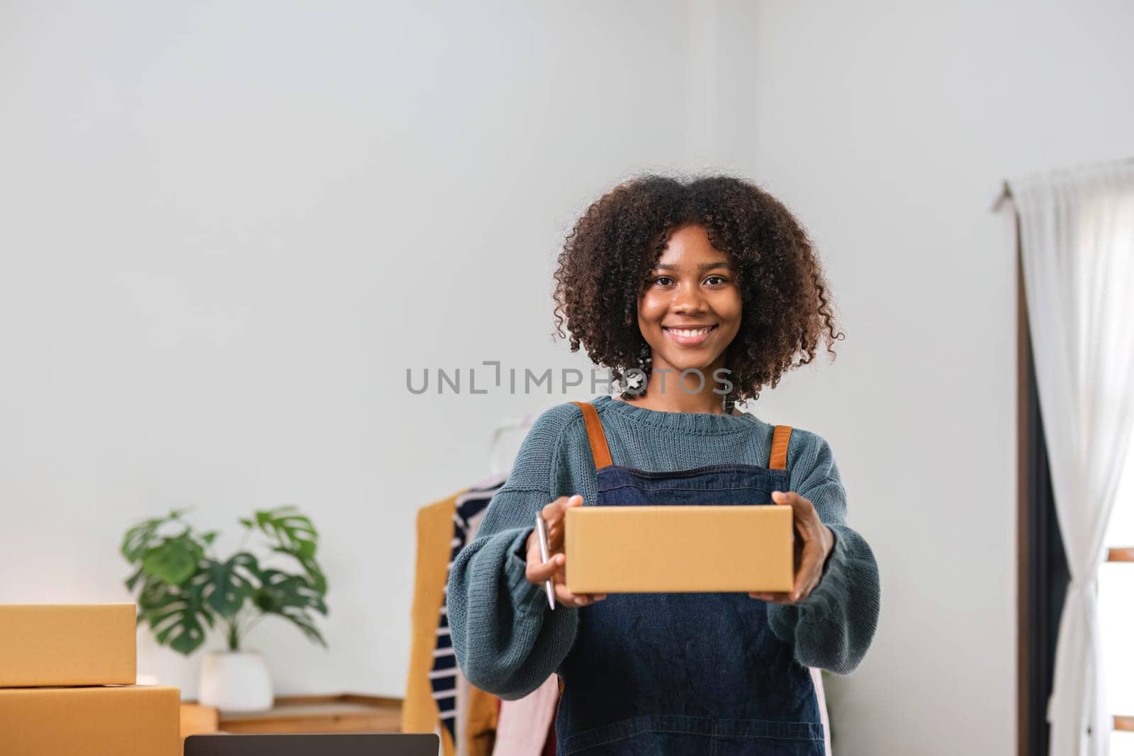 Young Asian woman business owner with many parcel boxes on the table happy online sales job, use your laptop, get an order from customers, take notes, and make arrangements for delivery by post...