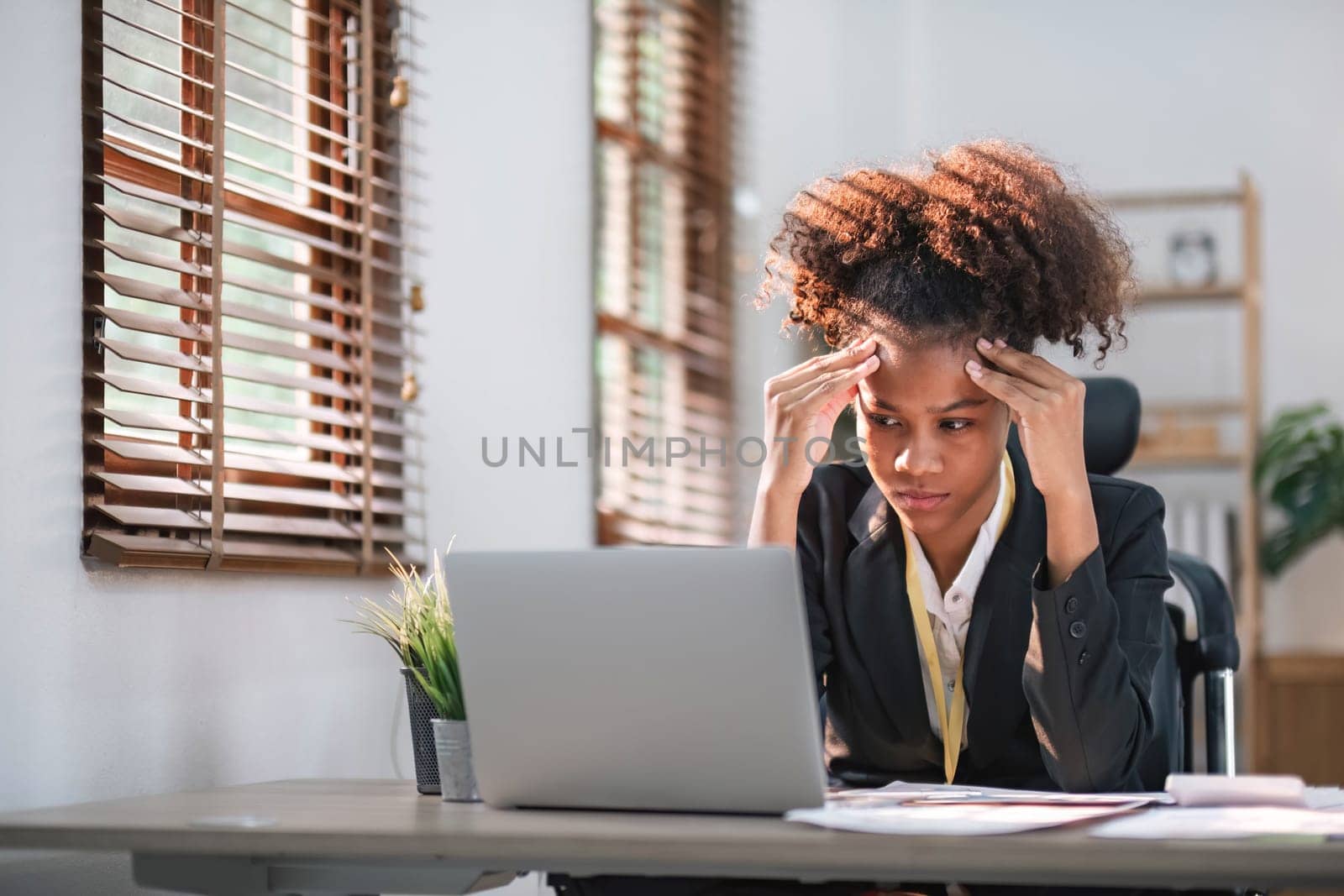 Young African American woman with afro hairstyle looks annoyed and stressed, sitting at the desk, using a laptop, thinking and looking at the camera, feeling tired and bored with depression problems..