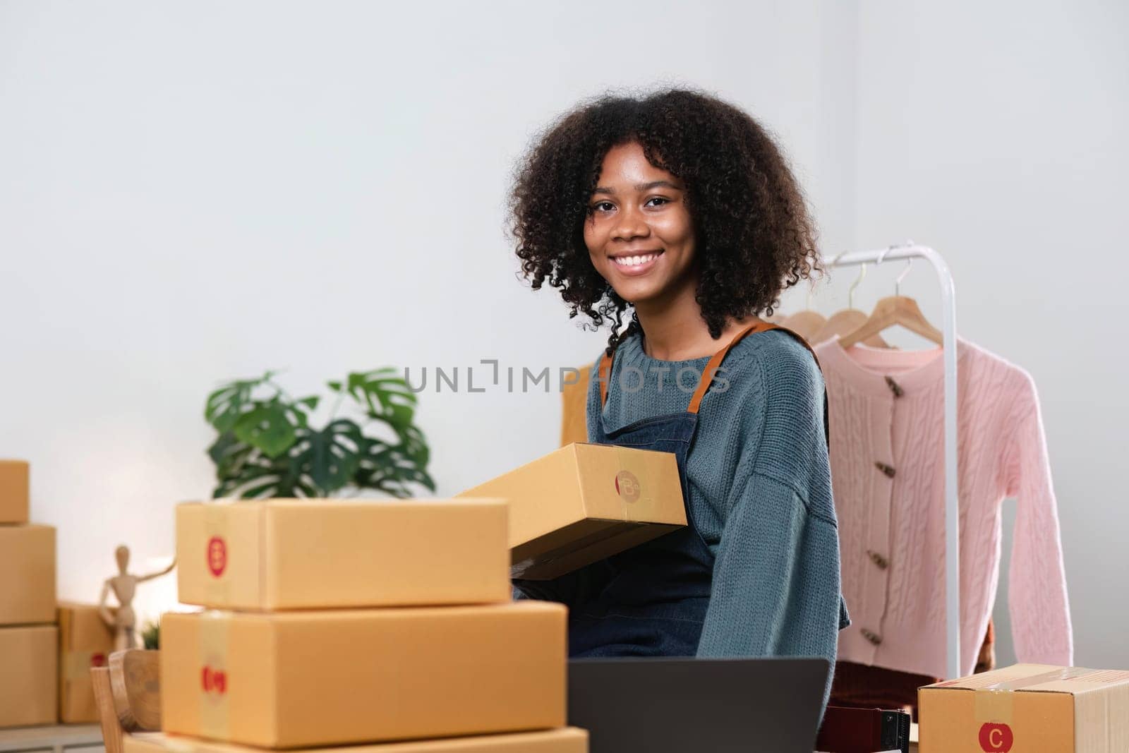 Young Asian woman business owner with many parcel boxes on the table happy online sales job, use your laptop, get an order from customers, take notes, and make arrangements for delivery by post...