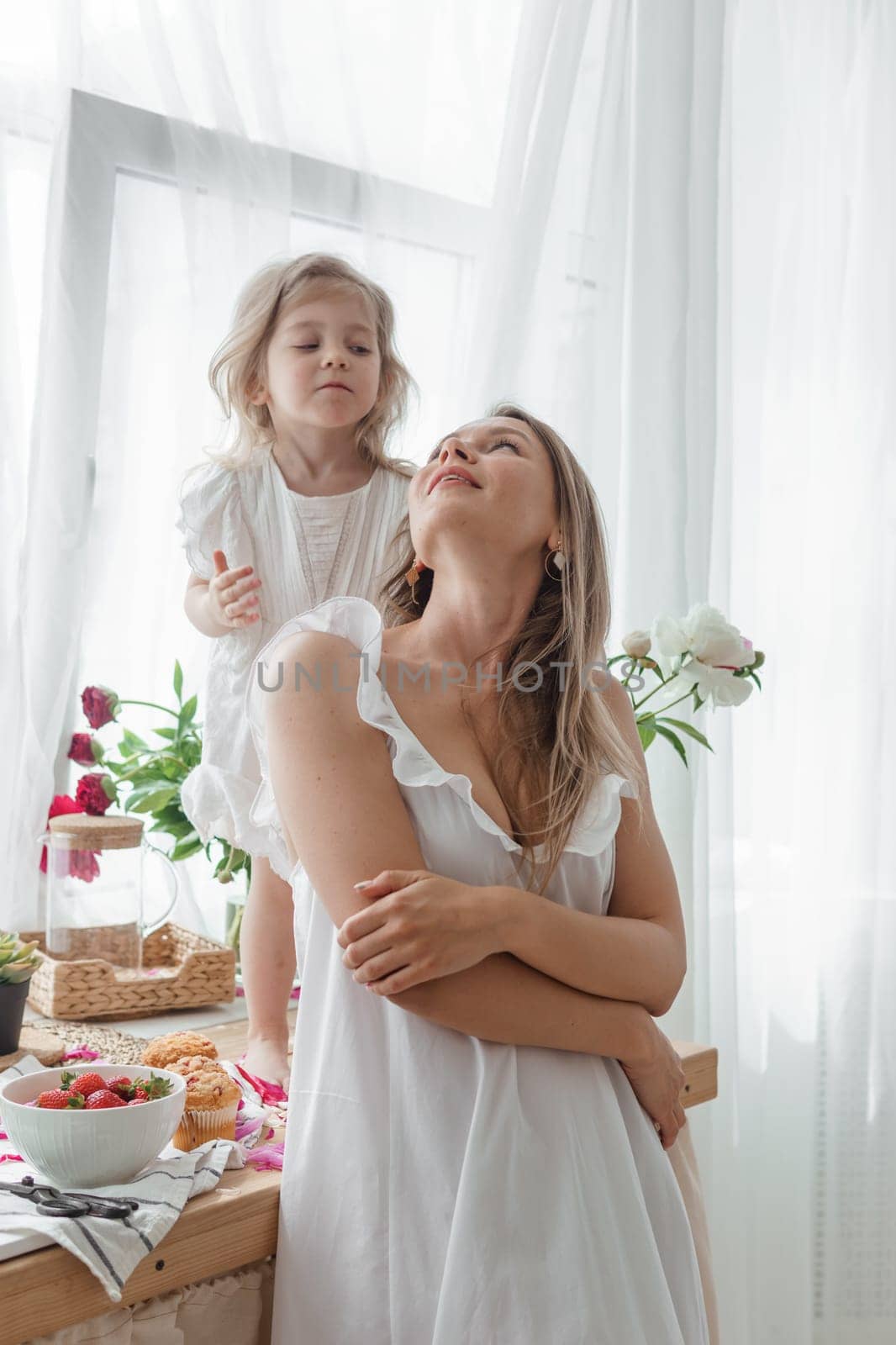 A little blonde girl with her mom on a kitchen countertop decorated with peonies. The concept of the relationship between mother and daughter. Spring atmosphere. by Annu1tochka