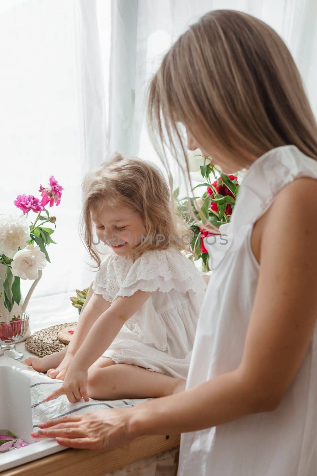 A little blonde girl with her mom on a kitchen countertop decorated with peonies. The concept of the relationship between mother and daughter. Spring atmosphere