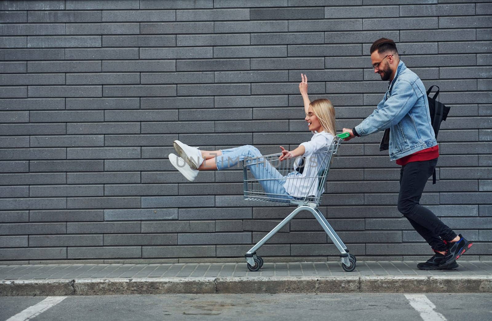 Having fun and riding shopping cart. Young stylish man with woman in casual clothes outdoors together. Conception of friendship or relationships.