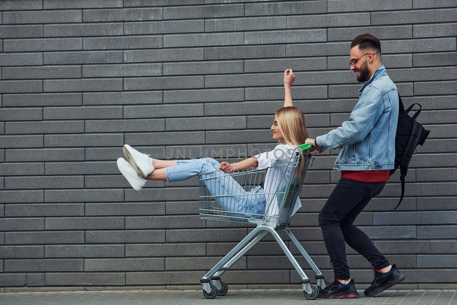 Having fun and riding shopping cart. Young stylish man with woman in casual clothes outdoors together. Conception of friendship or relationships.