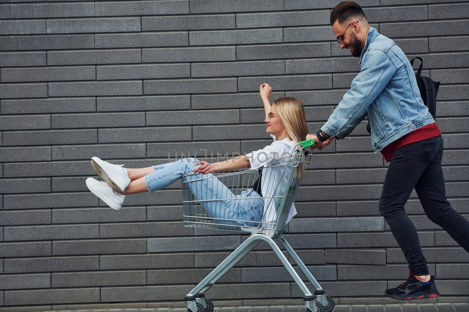 Having fun and riding shopping cart. Young stylish man with woman in casual clothes outdoors together. Conception of friendship or relationships.