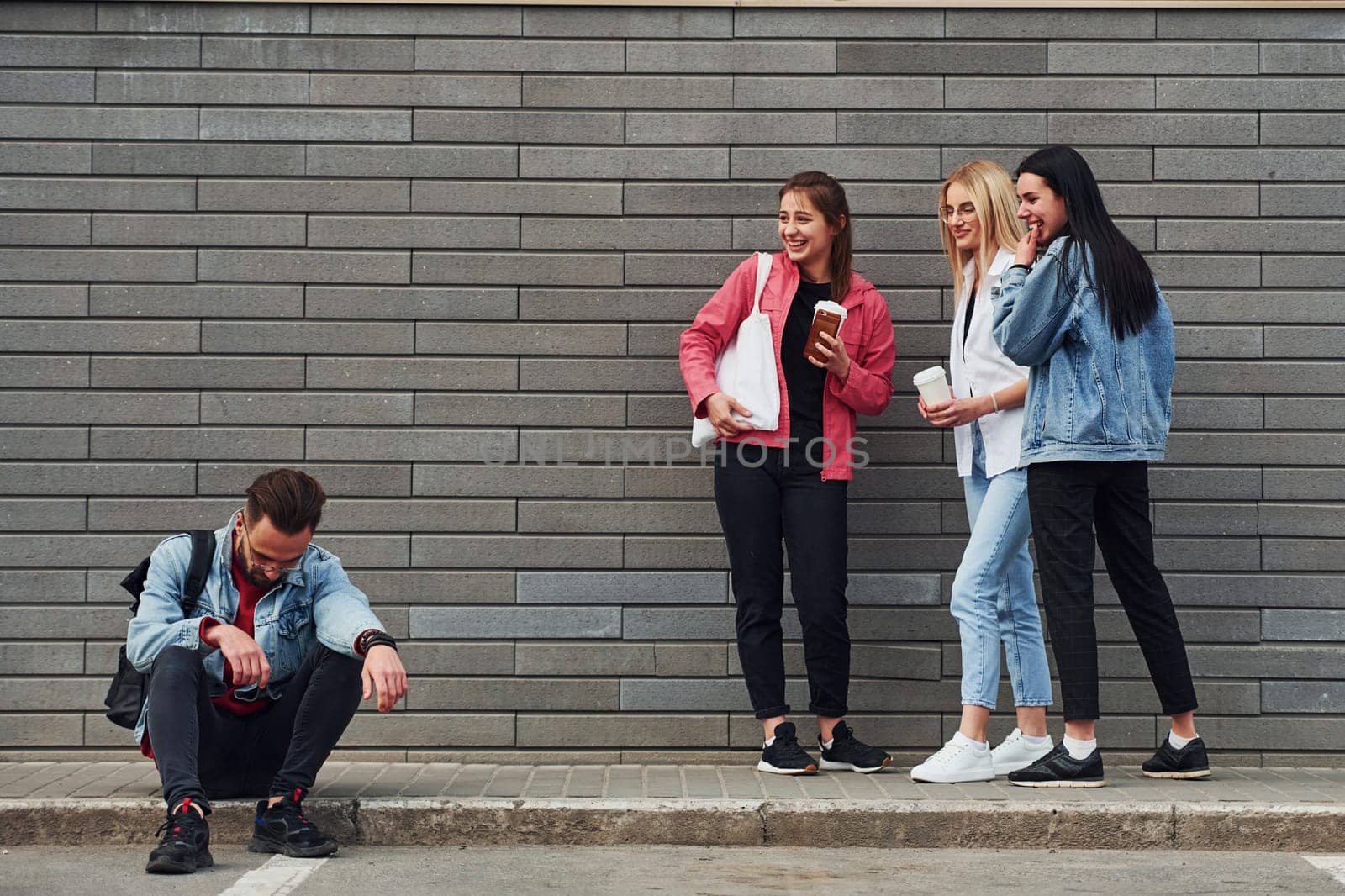 Three women standing and laughing at guy that sitting outdoors near building at daytime by Standret