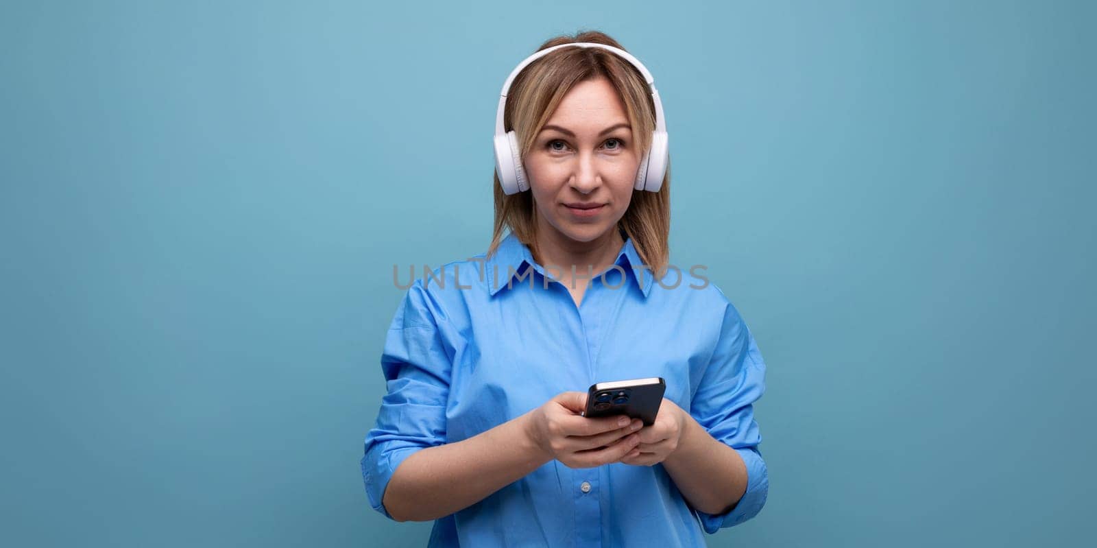 close-up of a millennial woman in a casual shirt with big white headphones with a smartphone in her hands on a blue isolated background with copy space by TRMK