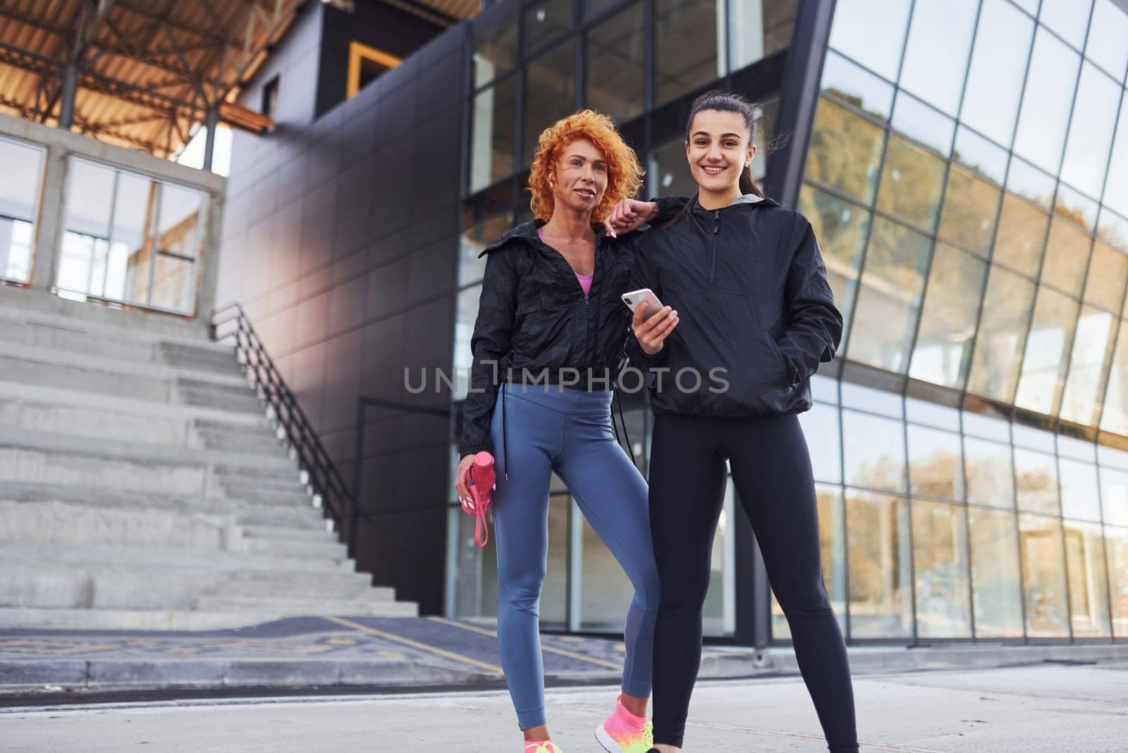 Holds phone in hands. Two female friends with sportive bodies have fitness day outdoors.