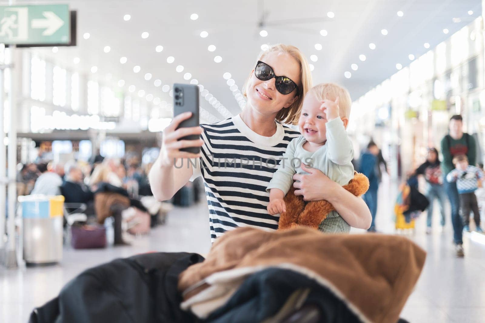 Mother taking selfie with mobile phone, while traveling with child, holding his infant baby boy at airport, waiting to board a plane. Travel with kids concept