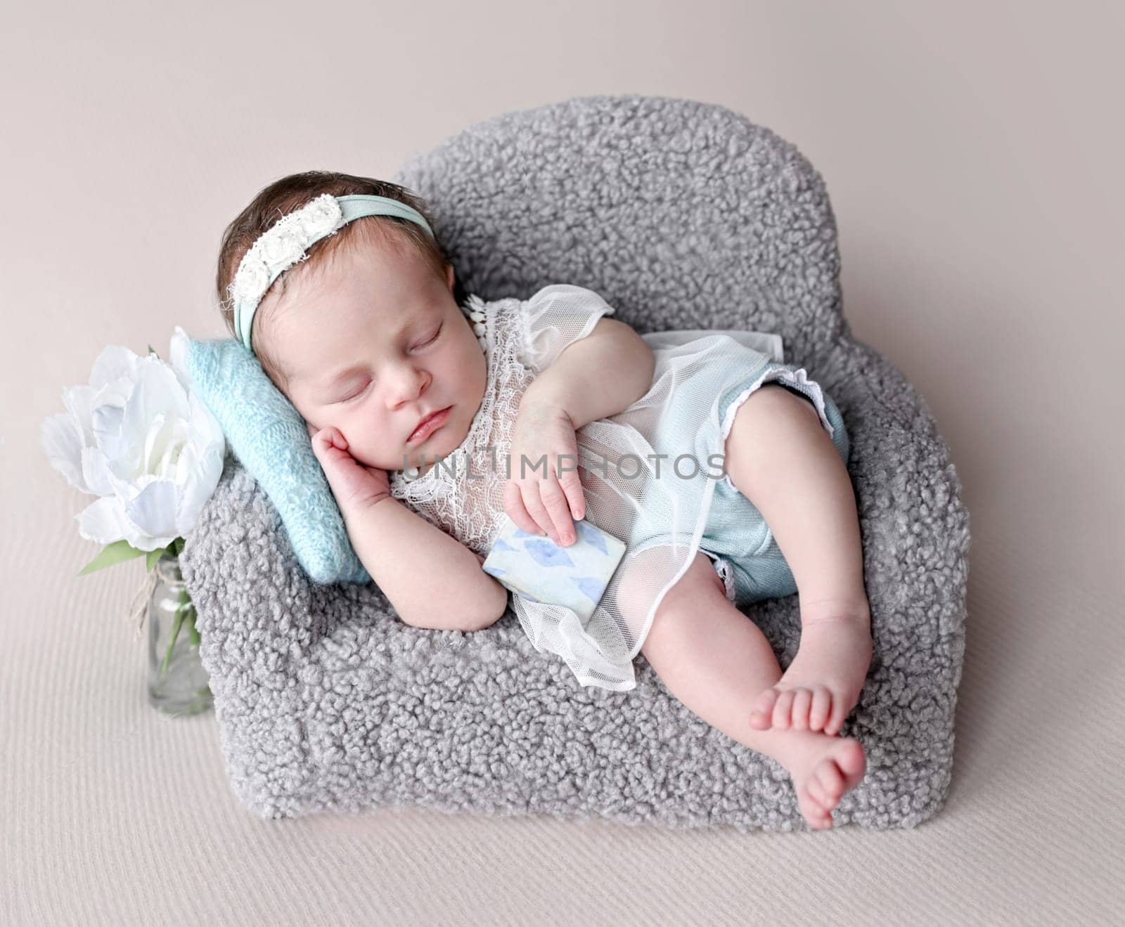 Newborn baby girl wearing dress sleeping on tiny sofa with flower decor. Cute infant child kid napping on couch