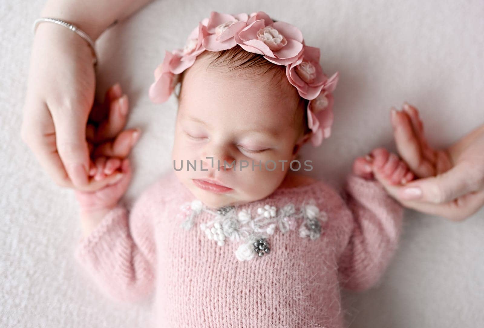 Newborn baby girl wearing flower wreath sleeping and mother holding her hands. Infant child kid napping, parent care and love