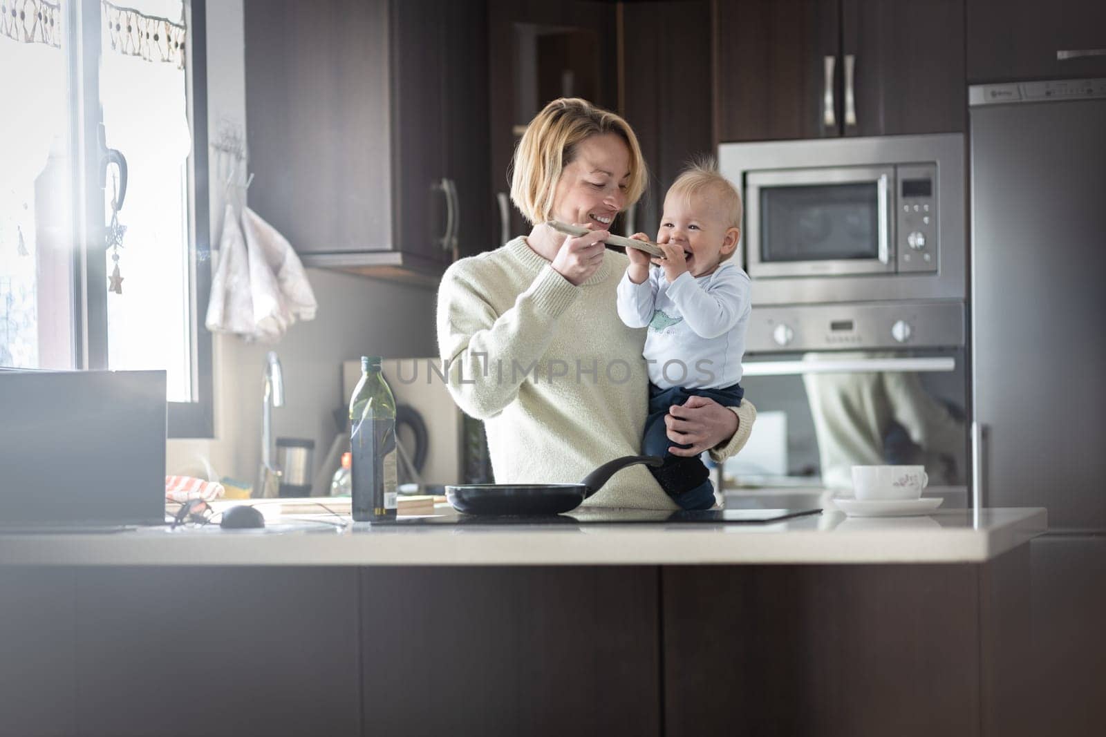 Happy mother and little infant baby boy cooking and tasting healthy dinner in domestic kitchen. Family, lifestyle, domestic life, food, healthy eating and people concept