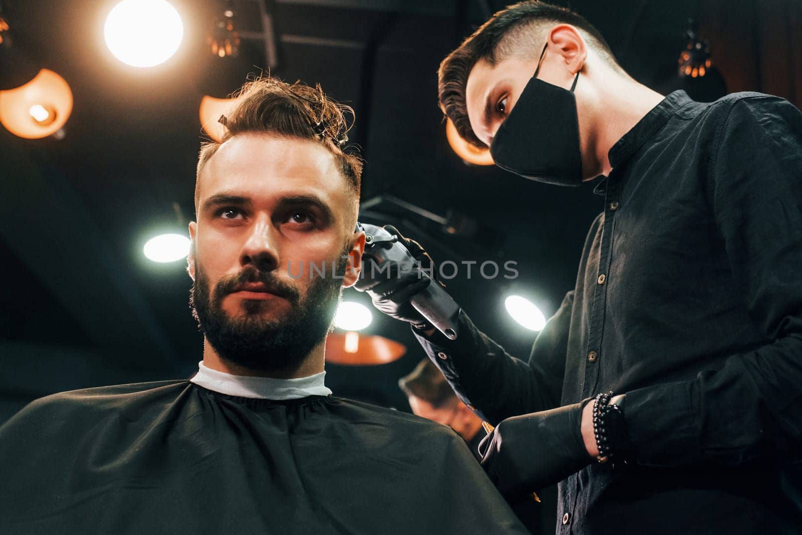 Front view of young bearded man that sitting and getting haircut in barber shop by guy in black protective mask.