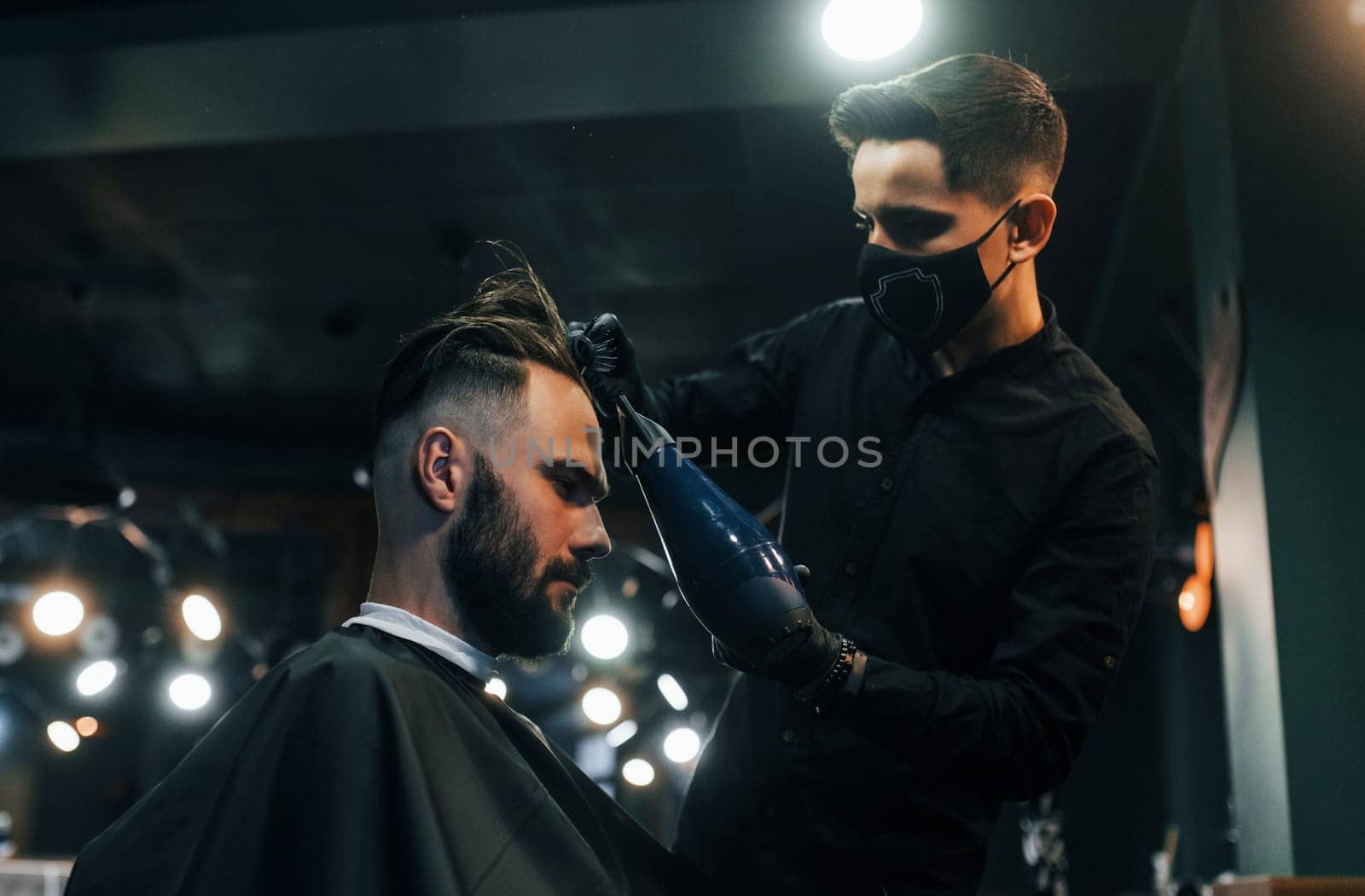Young bearded man sitting and getting haircut in barber shop by guy in black protective mask by Standret