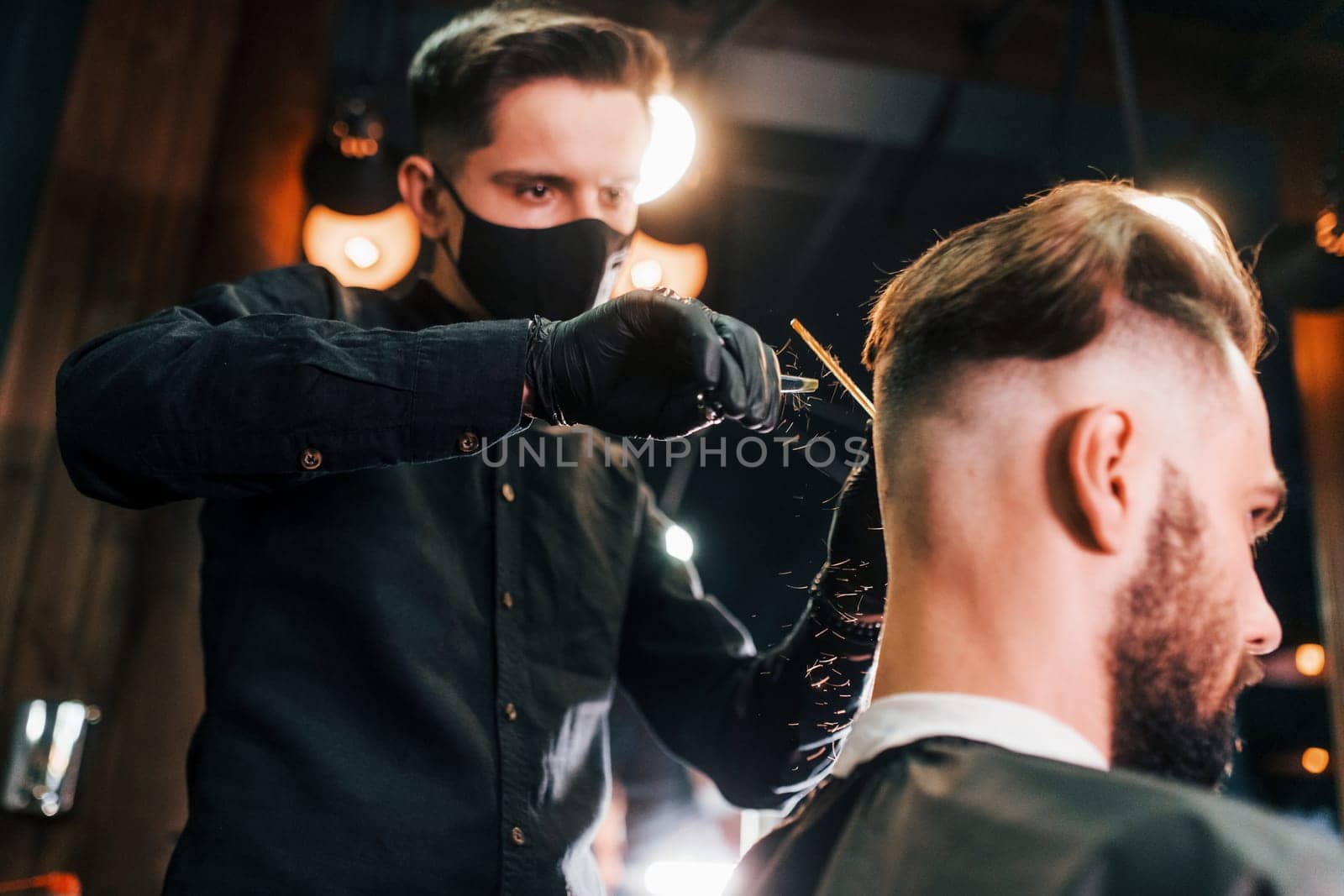 Young bearded man sitting and getting haircut in barber shop by guy in black protective mask.
