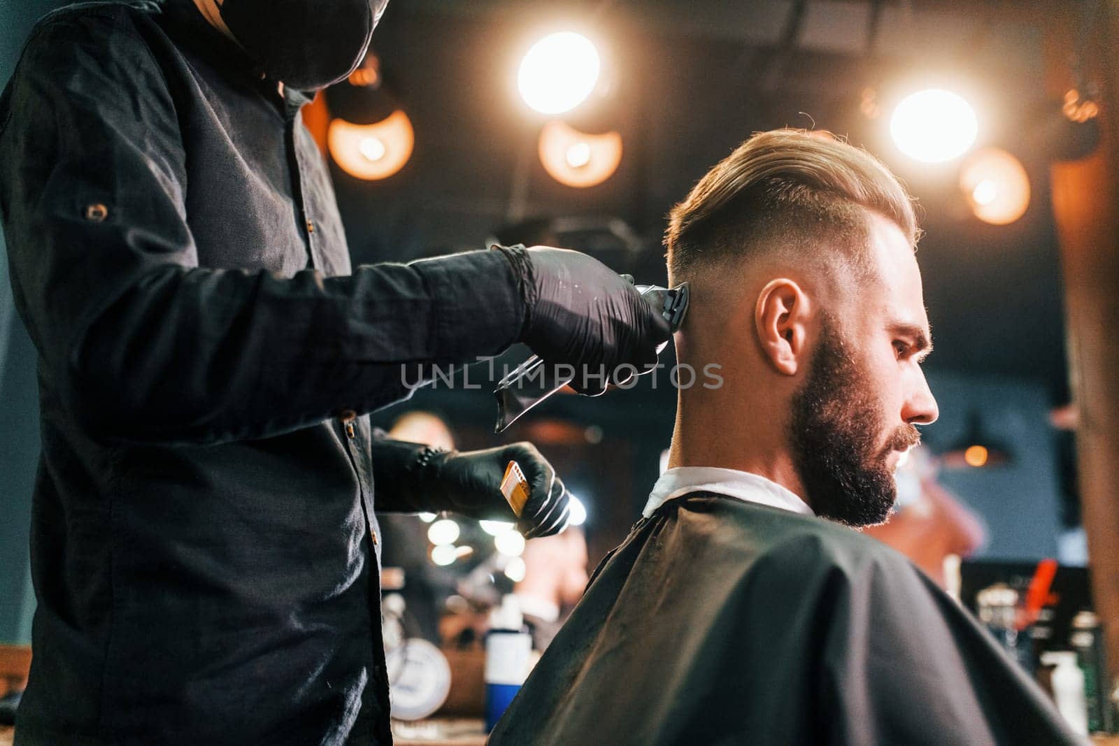 Young bearded man sitting and getting haircut in barber shop by guy in black protective mask.