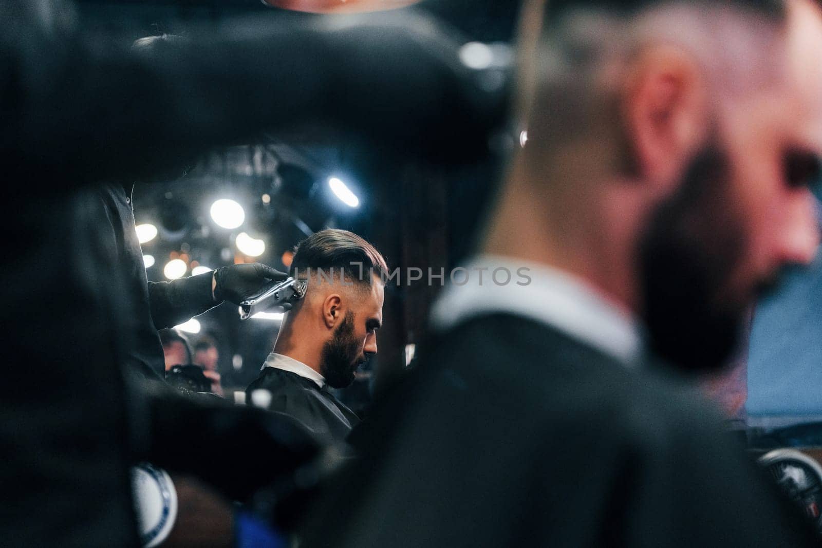 Young bearded man sitting and getting haircut in barber shop by Standret