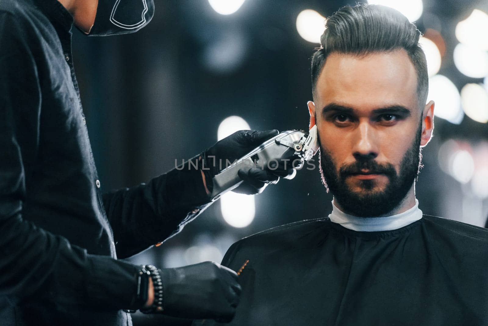 Young man with stylish hairstyle sitting and getting his beard shaved in barber shop by Standret
