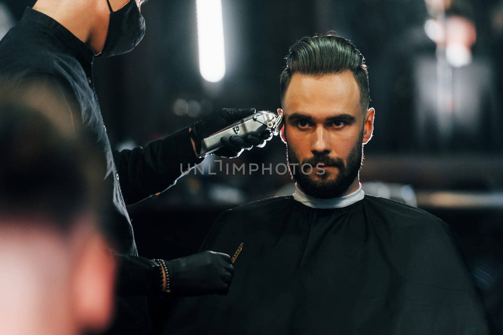 Young man with stylish hairstyle sitting and getting his beard shaved in barber shop.