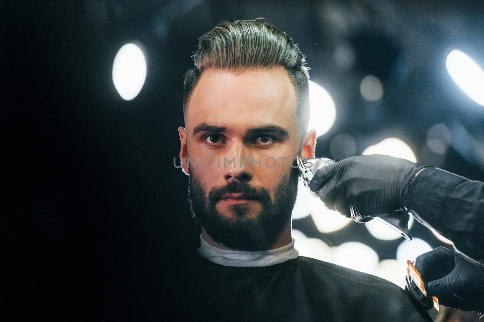 Young man with stylish hairstyle sitting and getting his beard shaved in barber shop.