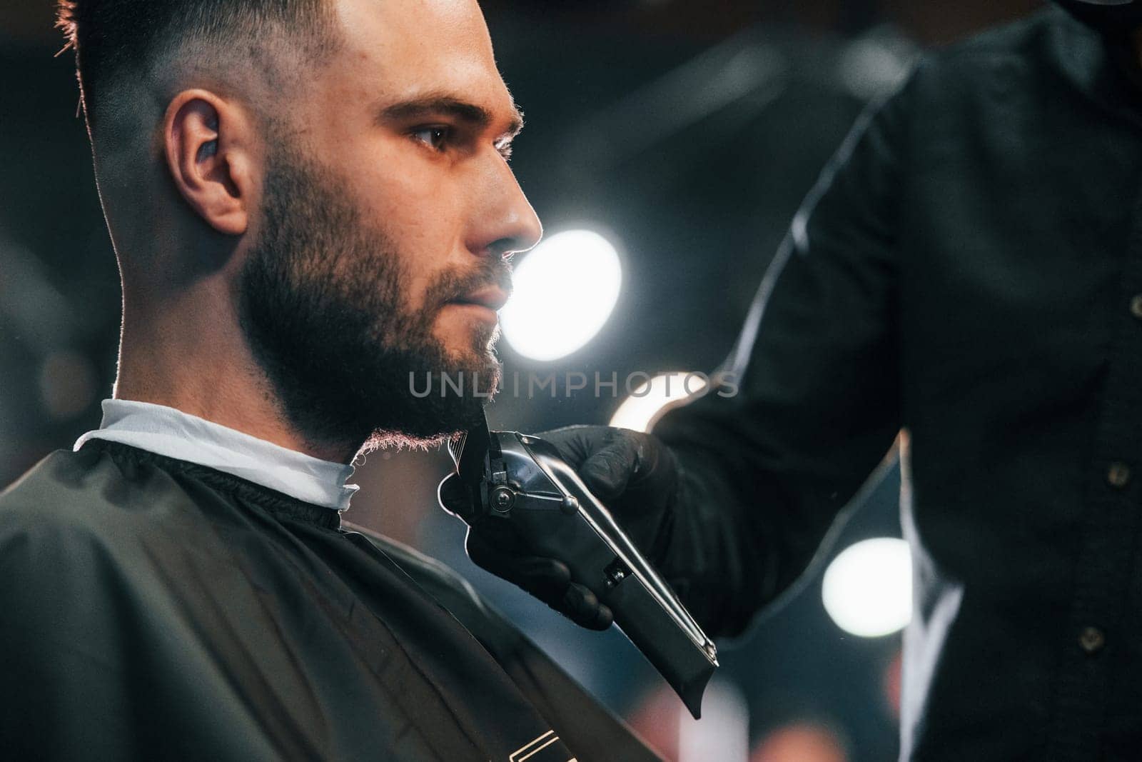 Young man with stylish hairstyle sitting and getting his beard shaved in barber shop.