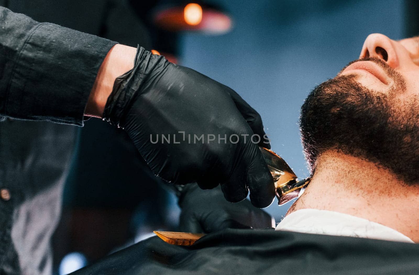 Young man with stylish hairstyle sitting and getting his beard shaved in barber shop by Standret