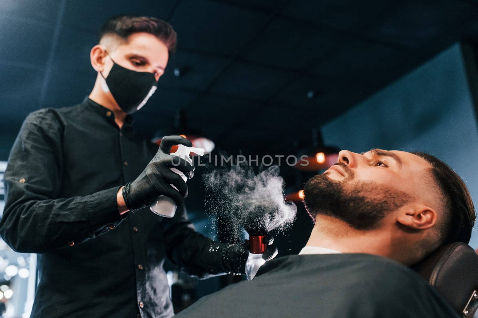 Young man with stylish hairstyle sitting and getting his beard shaved by guy in black protective mask in barber shop by Standret