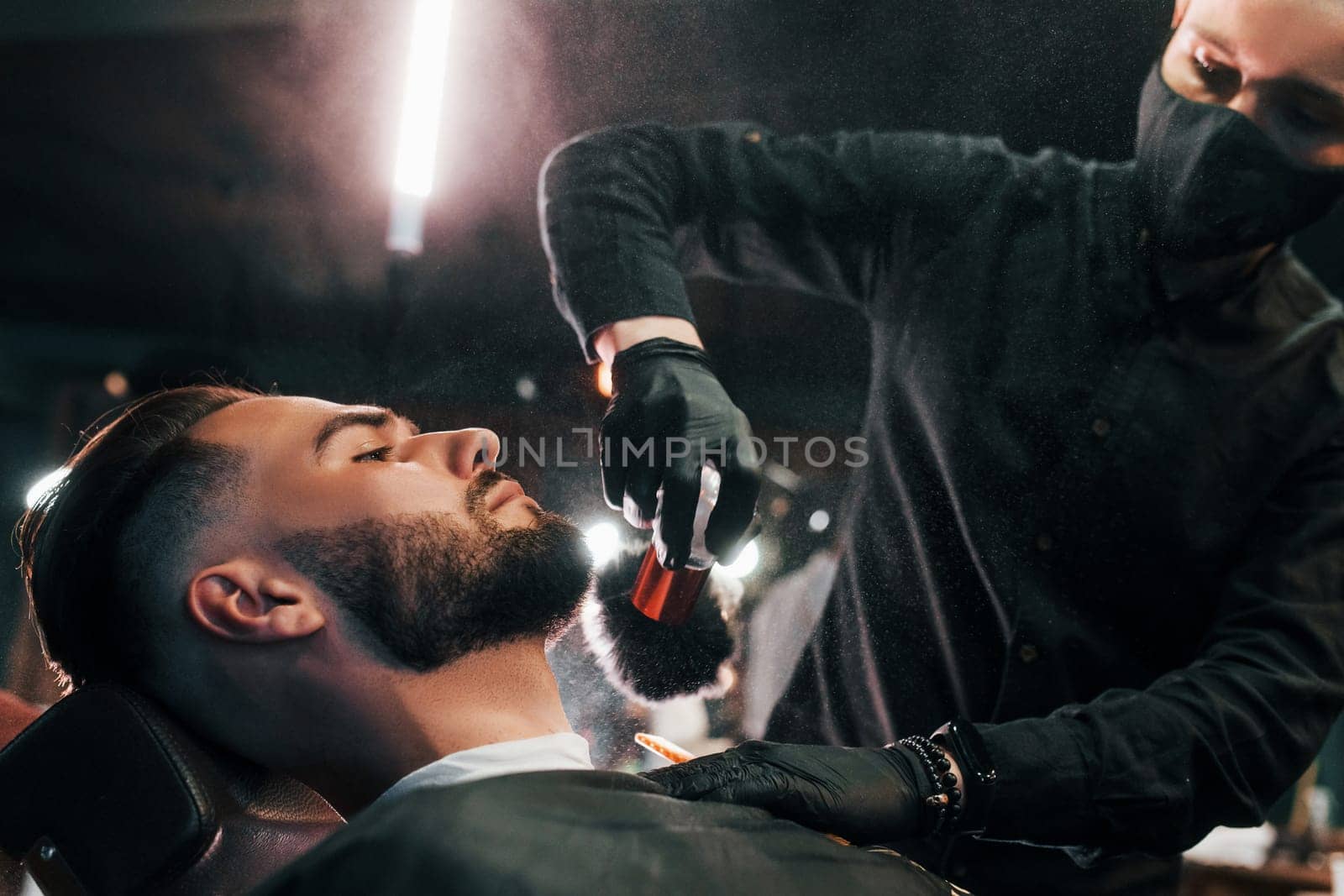 Young man with stylish hairstyle sitting and getting his beard shaved by guy in black protective mask in barber shop.