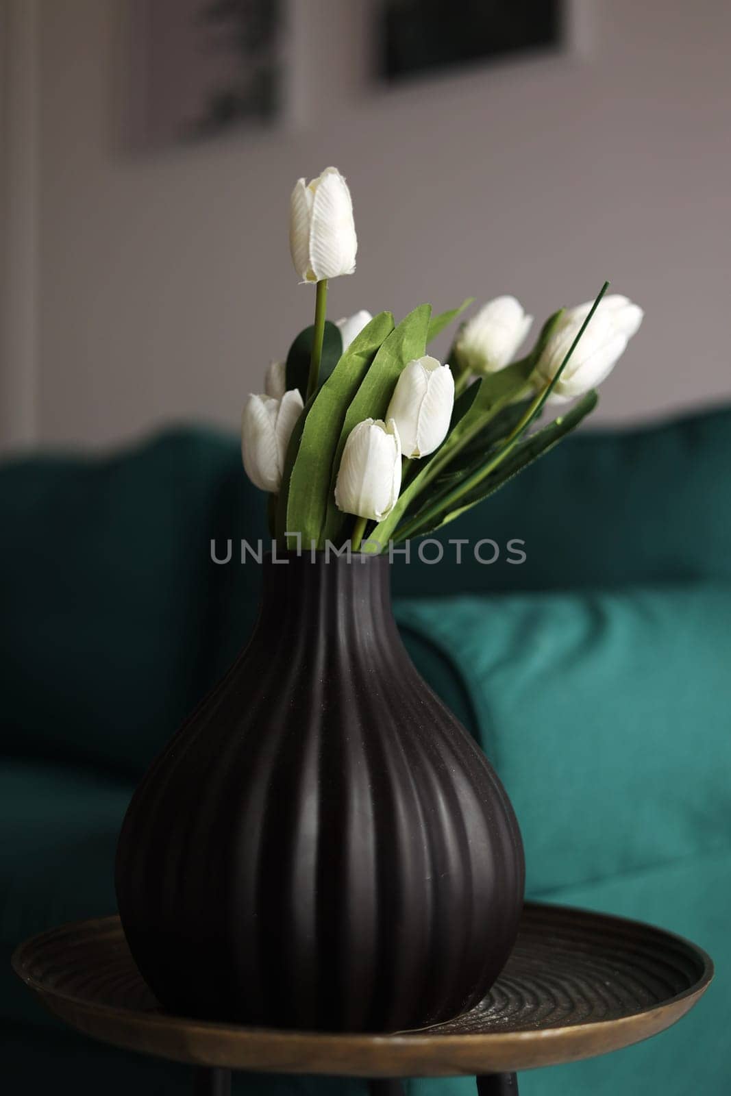 artificial white tulips and green leaves in a black vase on the table in home interior