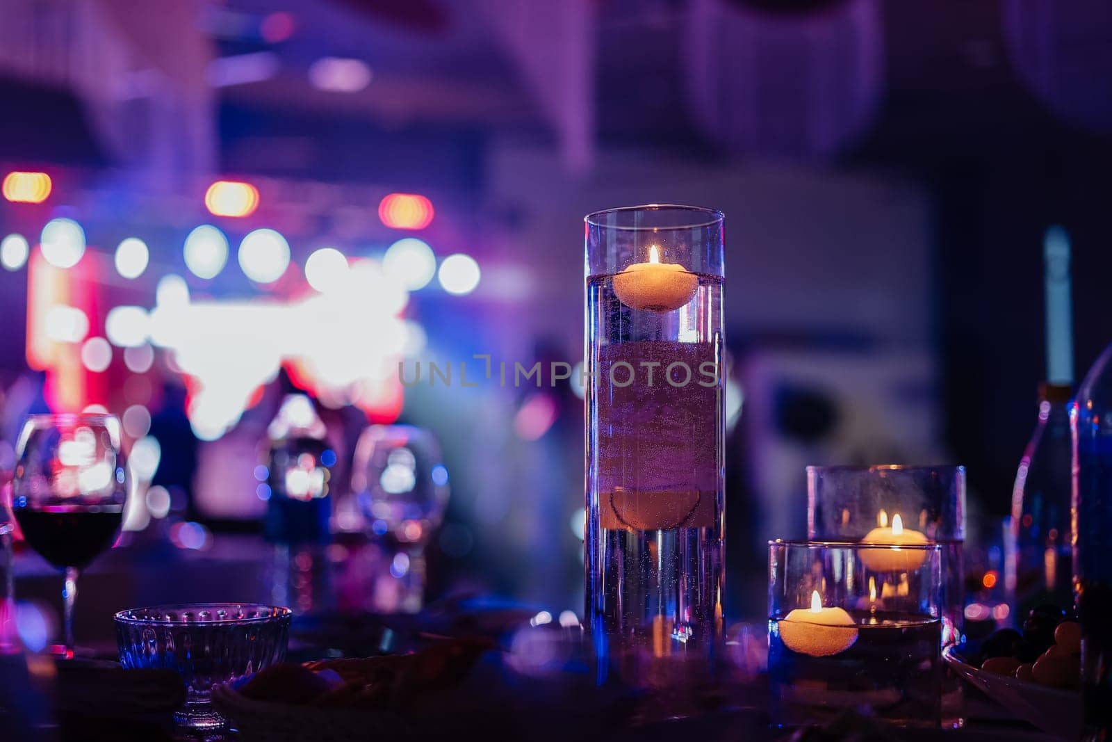 Banquet table decorated with burning candles in glass vases in restaurant hall. In the background party with silhouettes of people dancing on the dance floor with disco lights glowing searchlight. by Andriimedvediuk