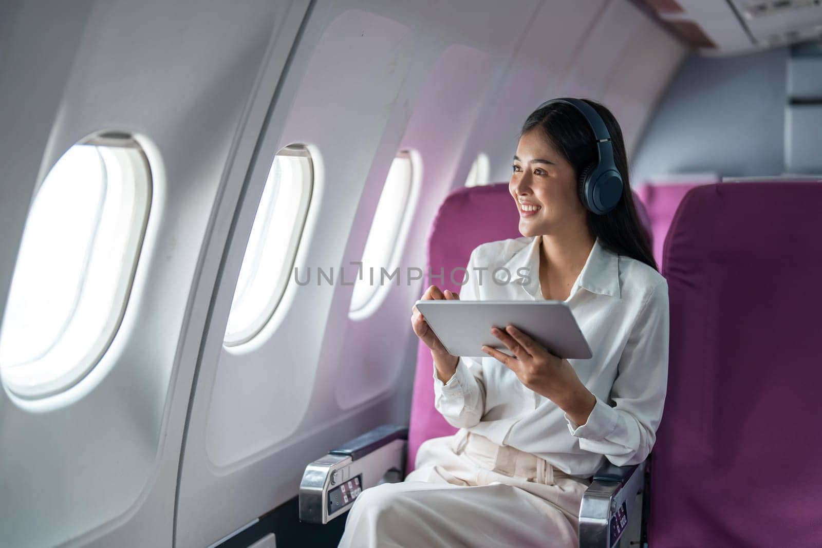 Young Asian business woman or female passenger wearing wireless headphone and working with tablet during the flight by nateemee