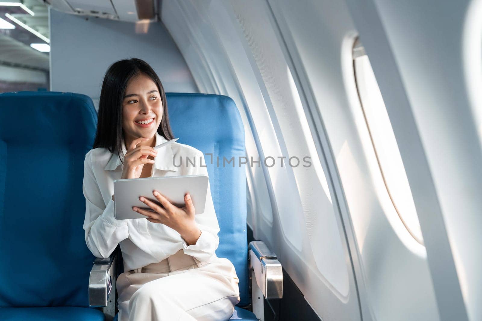 Young asian businesswoman in formal clothes working using tablet with smart pen while sitting in airplane cabin near window traveling to another place by nateemee