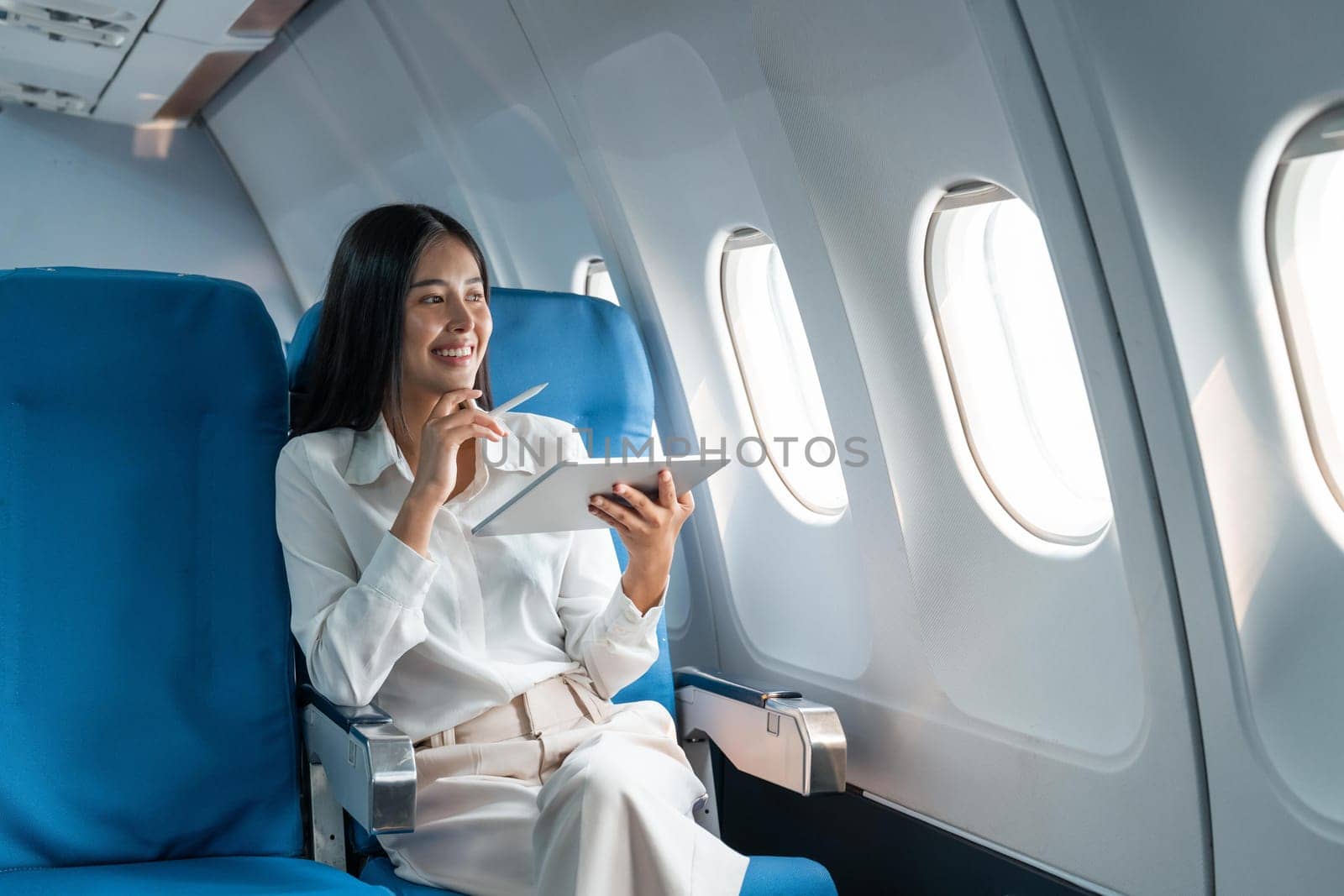 Young asian businesswoman in formal clothes working using tablet with smart pen while sitting in airplane cabin near window traveling to another place.