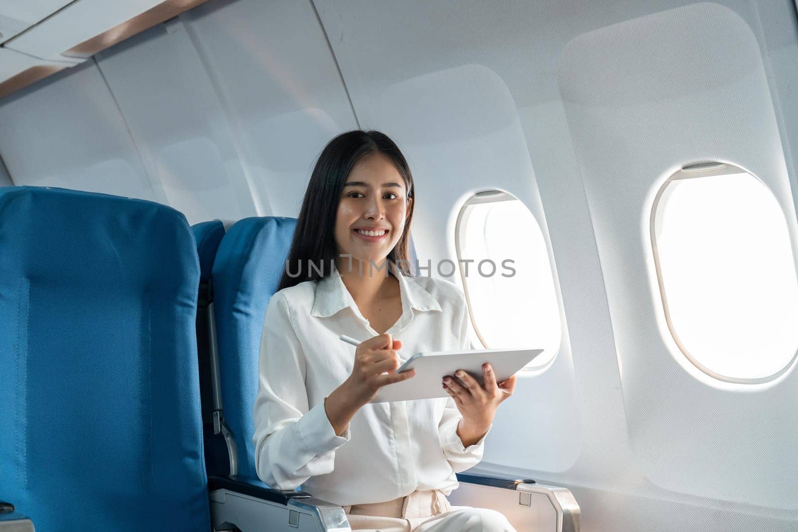 Young asian businesswoman in formal clothes working using tablet with smart pen while sitting in airplane cabin near window traveling to another place.