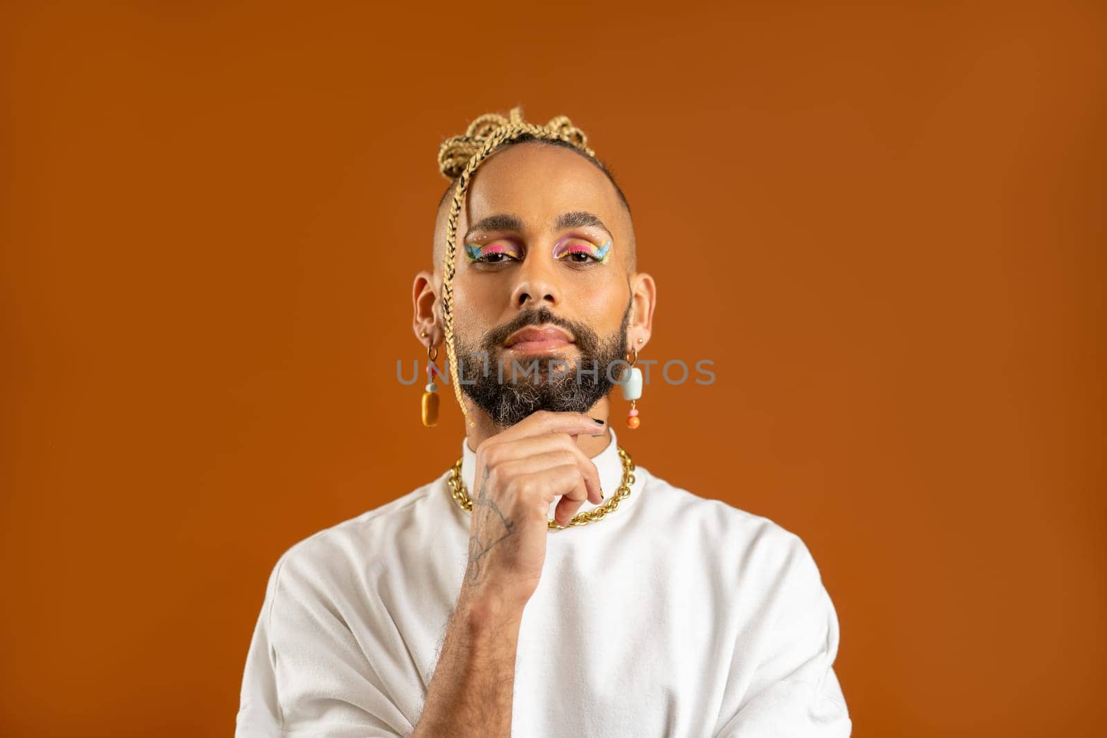Confident bearded black gay man with bright makeup standing isolated on orange background, dressed white. Exudes sense of pride and individuality. Diversity power of personal style