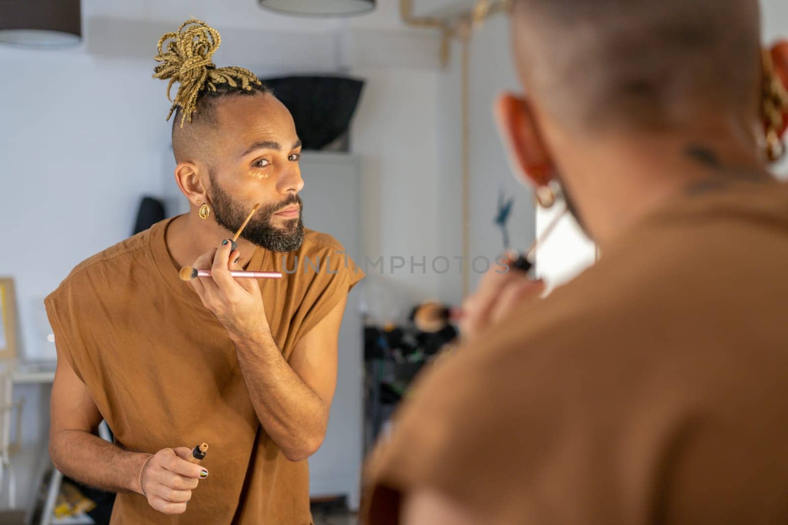 Black male gay applying make up looking mirror standing back to camera. Stylish homosexual man in dressing room doing make-up on face