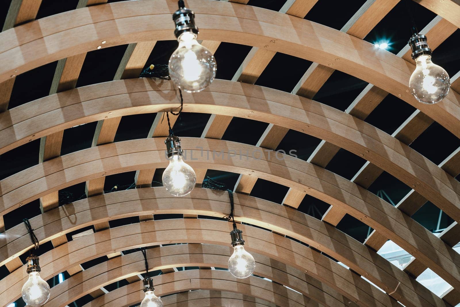 Many White Light Bulbs are hung on the Ceiling of the Wooden Curved Pergola Structure. Light Bulbs in Wooden Facade Architecture. Space for text, Selective focus.