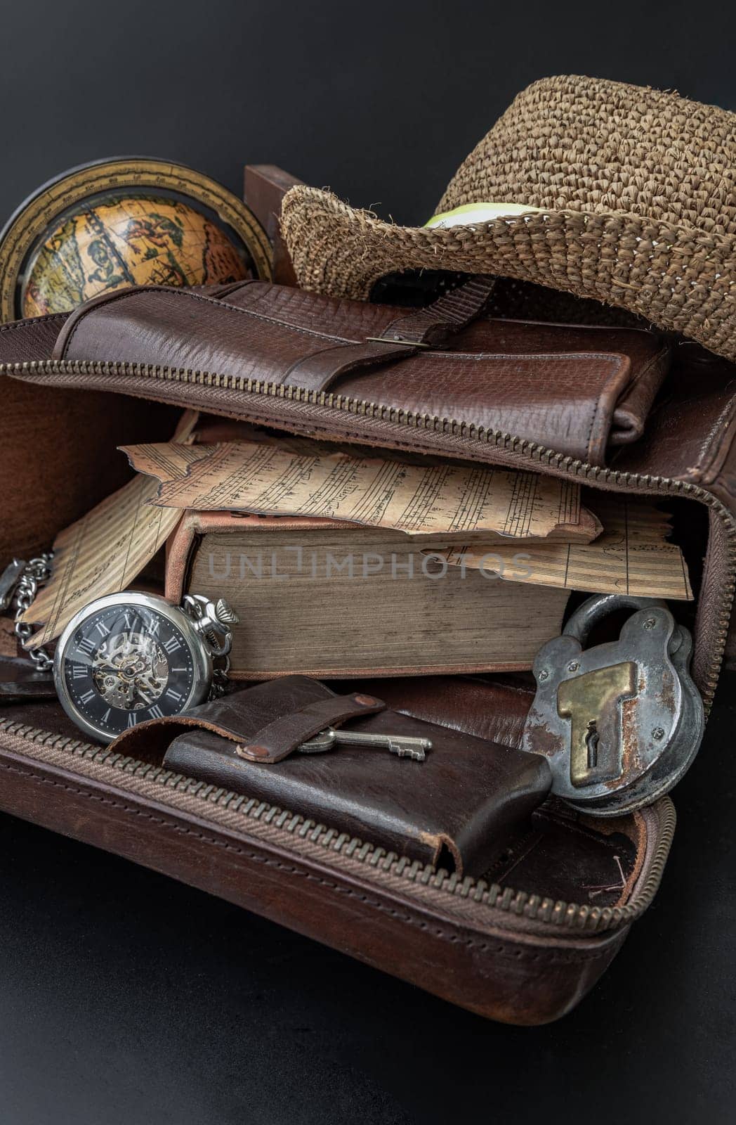 Bangkok,Thai - Mar 17, 2023 : The Vintage Music Notes, the Old Book, the Analog Pocket Watch, the Key and the Antique Iron Padlock are in the Old Brown Leather Bag. The Straw fedora Hat and the Globe are on them with Dark Background. The Good Memories are in Collectibles and Nostalgic Objects.